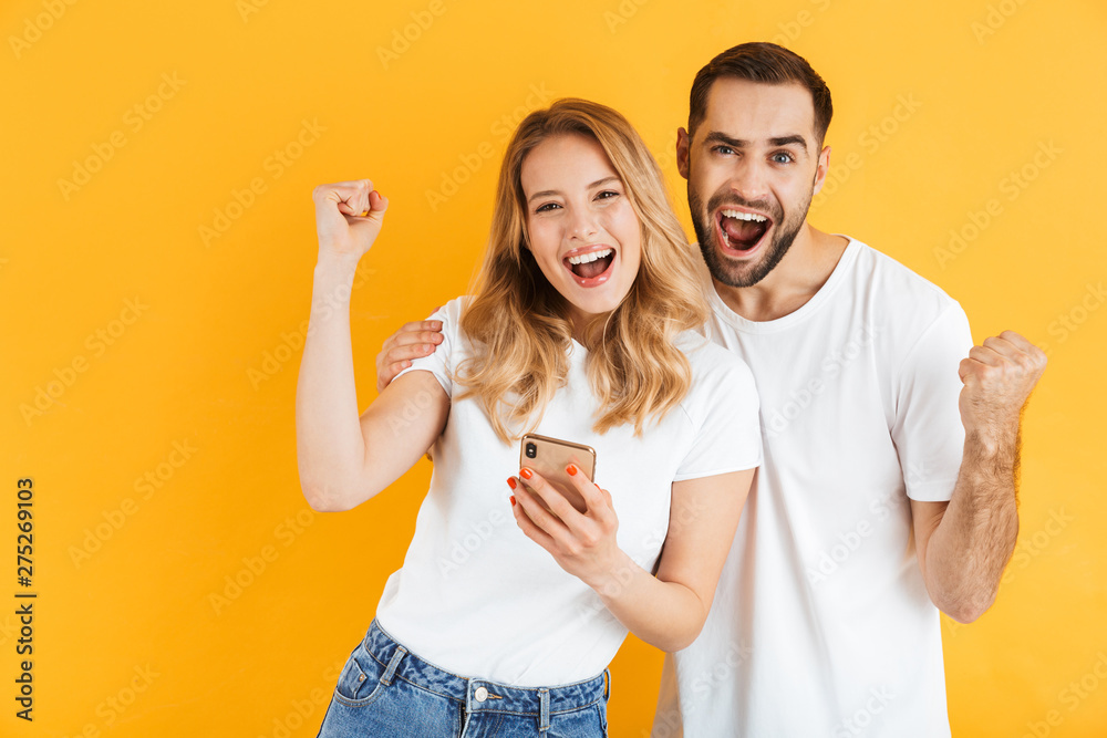 Portrait of young couple man and woman in basic t-shirts smiling and clenching fists while using cellphones together Portrait of young couple man and woman in basic t-shirts smiling and clenching fists while using cellphones together