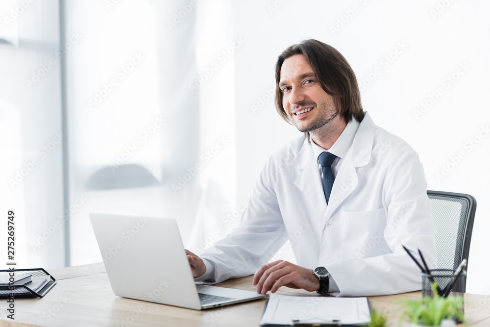 happy doctor in white coat looking at camera while working with laptop happy doctor in white coat looking at camera while working with laptop
