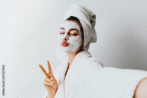 Self portrait of charming, stylish, pretty, model after bath wrapped in towel shooting selfie on front camera applying using face mask for her dry, oiled, sensual face skin, enjoying procedure photo