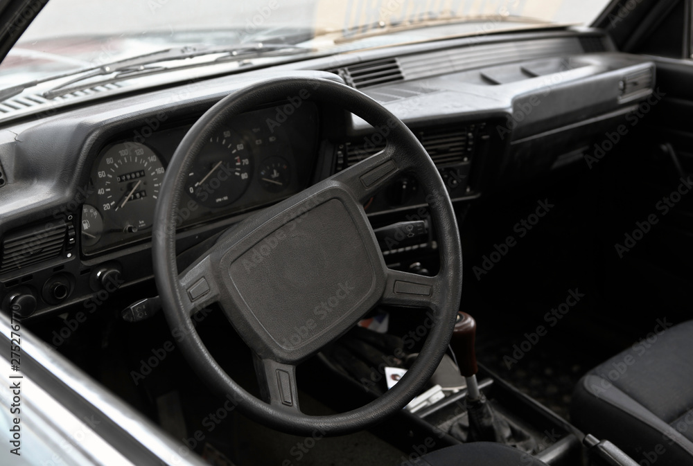 Old shcool car interior, steering wheel in focus, close up view. Gauges, sensors, gear shift knob, glove box blurred in the background. Black plastic, dark fabric cover.