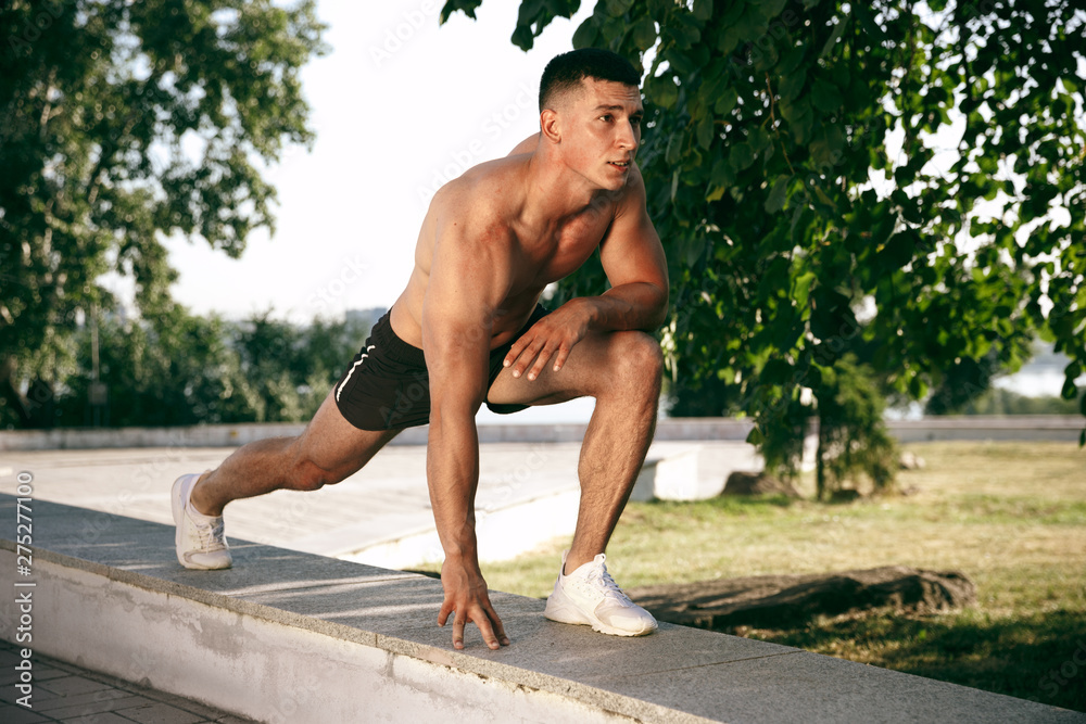 A muscular male athlete doing workout at the park. Gymnastics, training, fitness workout flexibility. Summer city in sunny day on background field. Active and healthy lifestyle, youth, bodybuilding.