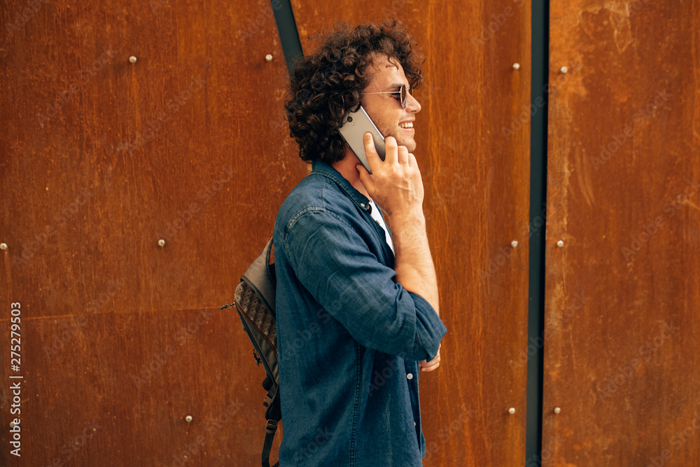 Rear view of happy man standing outdoors, typing messages on mobile phone. Young male with curly hair wears sunglasses walking in the city browsing on his cell phone on modern industrial wall.