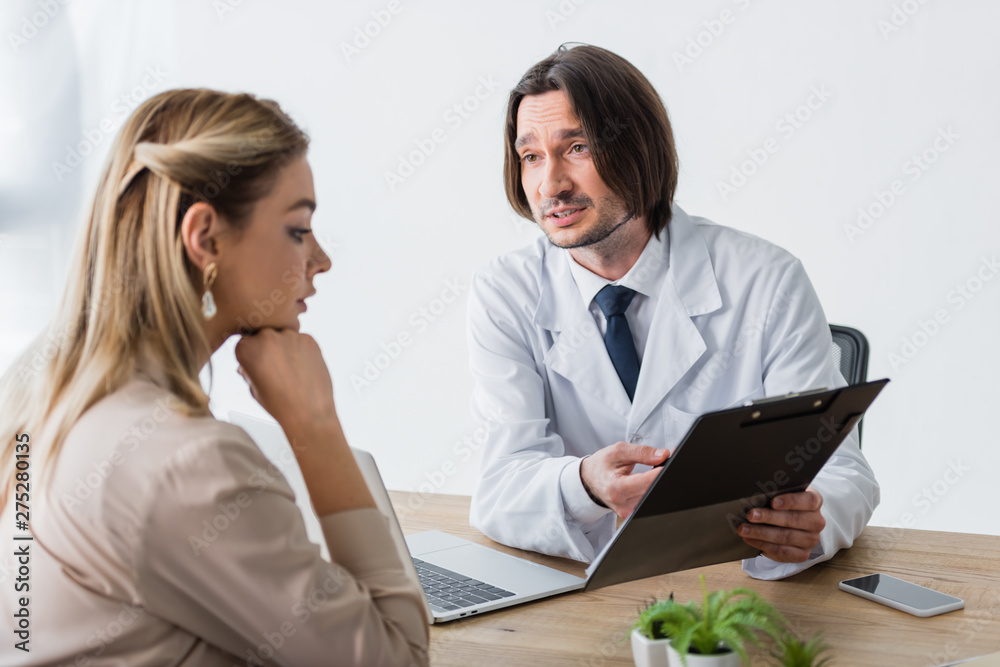 handsome doctor holding document in hands, looking at patient and sitting behind wooden table handsome doctor holding document in hands, looking at patient and sitting behind wooden table