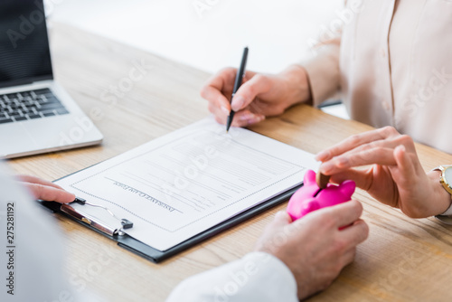 partial view of doctor holding piggy bank in hand while woman holding coin and signing document