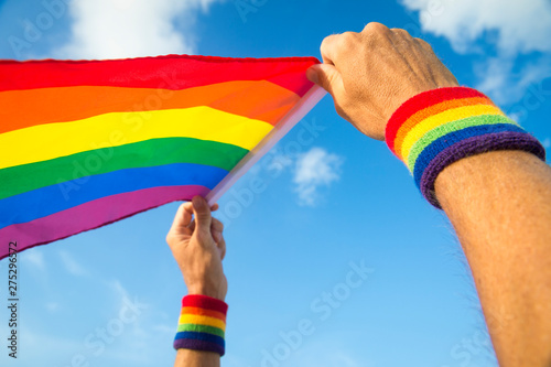 Hands with rainbow color wristbands waving gay pride flag backlit in the wind against a vibrant blue sky