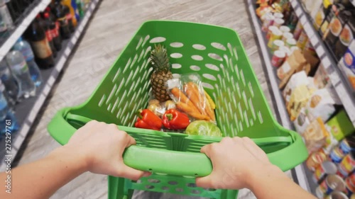 First-person view of female hands moving shopping trolley with fresh colorful vegetables and fruits through aisles of supermarket, sale and shopping concept