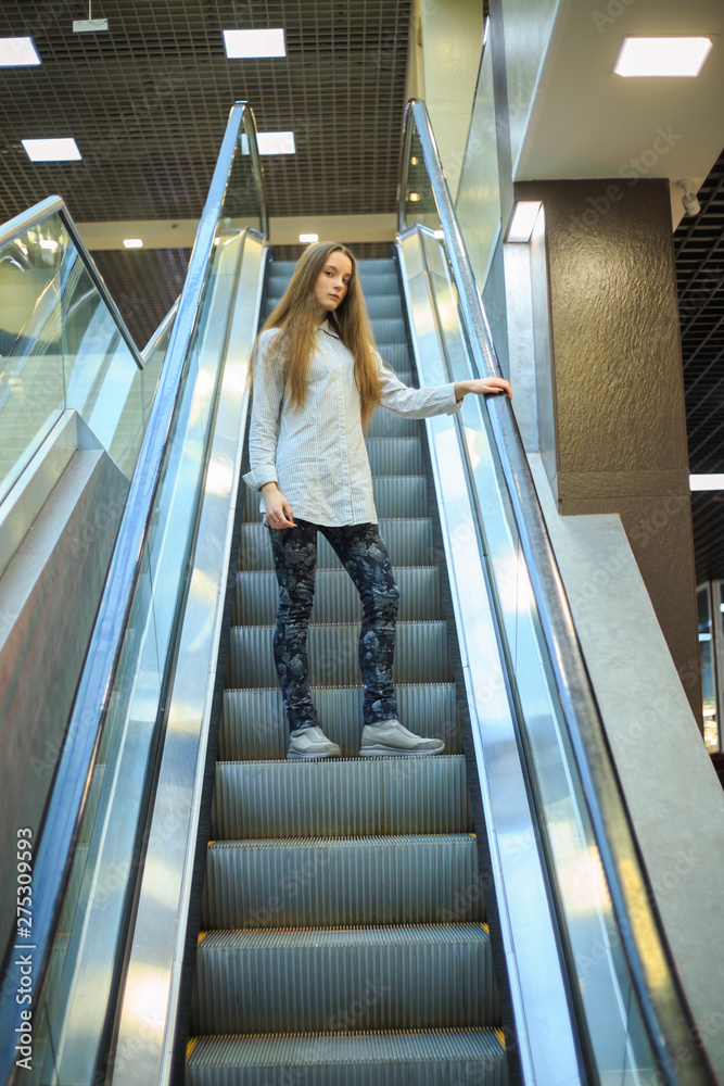 Portrait of a young pretty caucasian teenage girl with long hair in a shirt on the escalator in a mall.