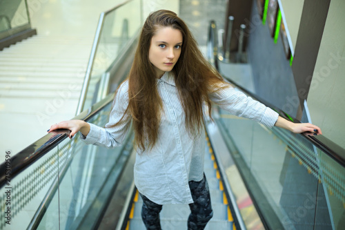 Portrait of a young pretty caucasian teenage girl with long hair in a shirt on the escalator in a mall. © aleksandr_yu