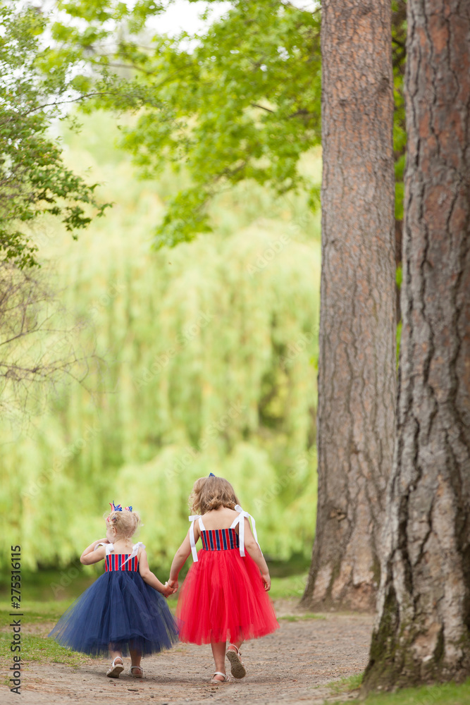 Two adorable young sisters wearing 4th of July dresses holding hands and walking down a park path Two adorable young sisters wearing 4th of July dresses holding hands and walking down a park path