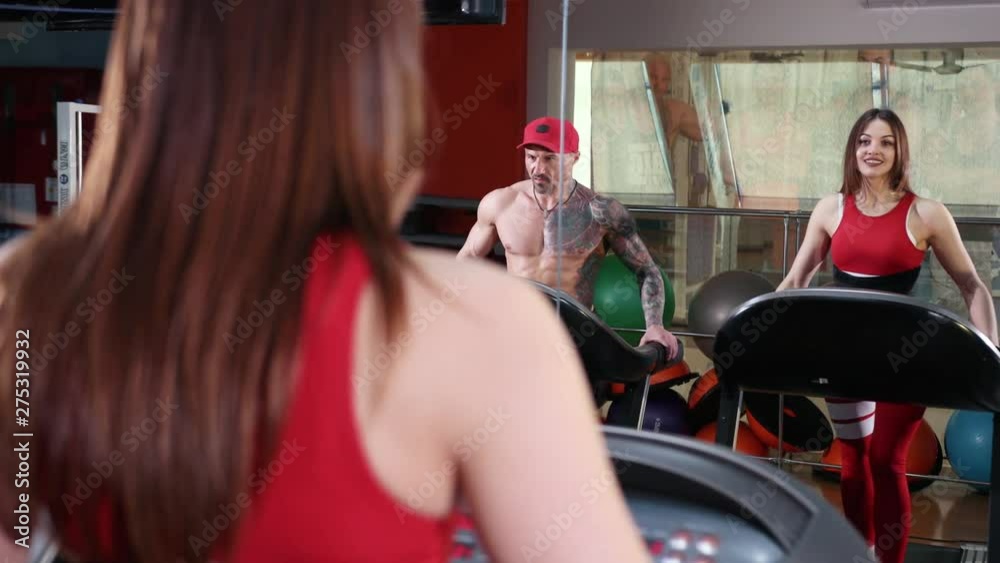 Attractive beautiful caucasian girl and muscular athletic man running on the treadmill in the sport gym. Woman in a red sports suit, and a bodybuilder in a cap. Close-up, Prores, Slow Motion Attractive beautiful caucasian girl and muscular athletic man running on the treadmill in the sport gym. Woman in a red sports suit, and a bodybuilder in a cap. Close-up, Prores, Slow Motion