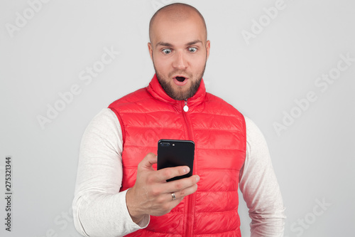Photo of stunned male reads text message with surprised expression, holds cell phone, finds out something shocking, connected to wireless internet. People and technology concept photo