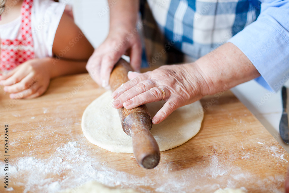 Hands of grandmother roll out dough with rolling pin and flour on wooden table. Family is cooking pastries in kitchen. Senior woman and child prepare homemade pie or pizza