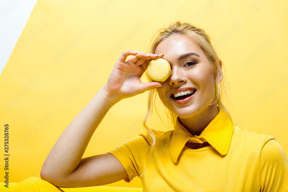 cheerful blonde girl smiling while covering face with tasty macaron on yellow cheerful blonde girl smiling while covering face with tasty macaron on yellow