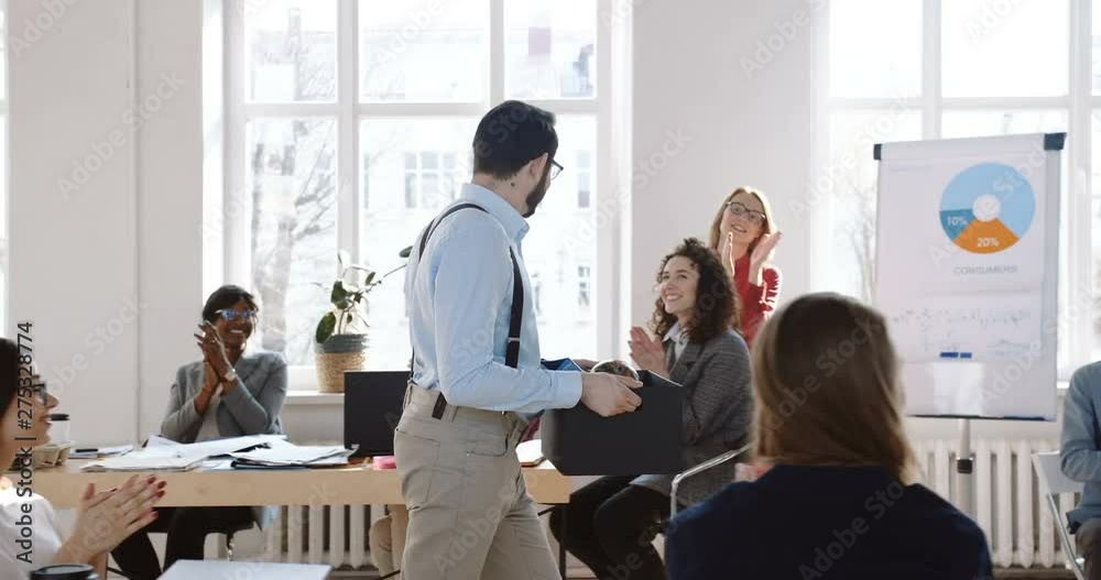 Dolly shot of happy smiling young male manager walking along modern office with stuff box, colleagues cheering him up. Dolly shot of happy smiling young male manager walking along modern office with stuff box, colleagues cheering him up.