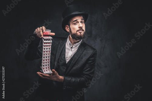 A Bearded magician in a black suit and top hat, showing trick with playing cards on a dark background.