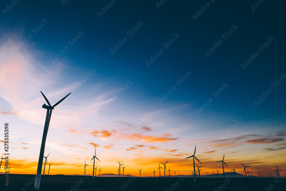 Silhouette of windmills station with propellers generating alternative clean green power from eco resources in rural agri environment against blue sunset sky. Farm of turbines producing electricity