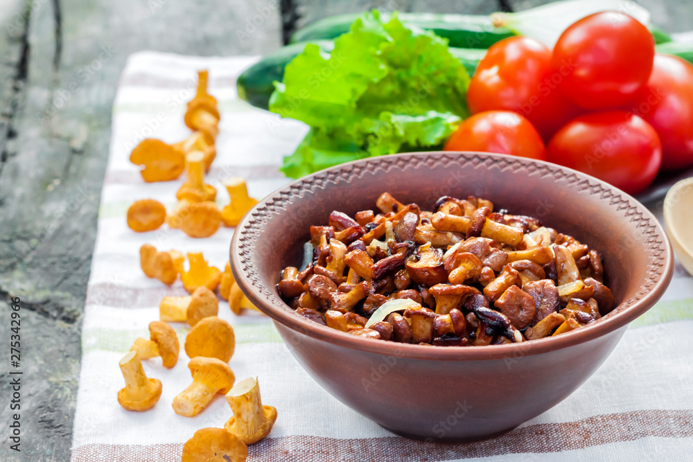 Fried chanterelles with onion in rustic bowl and plate with fresh vegetables for salad on background
