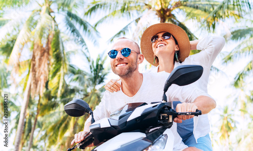 Happy smiling couple travelers riding motorbike during their tropical vacation under palm trees