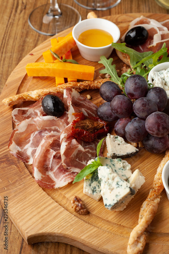 Close up of antipasto with assortment of cold snacks to wine: cheddar cheese, blue cheese, prosciutto, chorizo, olives, rucola, olive oil and pesto, served on a wooden dish