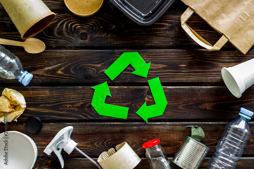 Green recycling sign with waste materials, paper bag and cup, plastic bottles, flatware on wooden background top view
