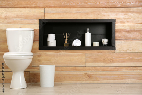 Toilet bowl at wooden wall with bathroom accessories in niche