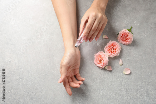 Woman applying rose essential oil on wrist and flowers at grey table, top view
