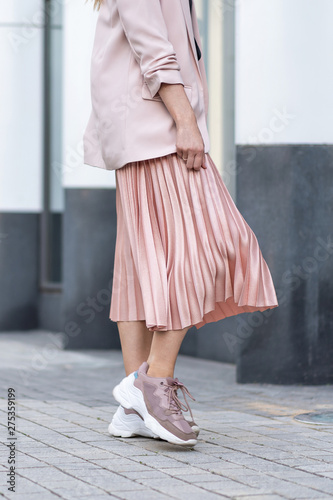 Model posing in a peach skirt, pleated, sneakers and jacket. Coral or pink color. On the street in the afternoon.