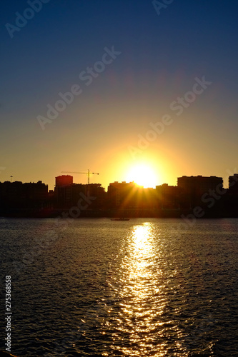 Seaside city skyline at sunset, residential buildings and cranes, shining water in the foreground.