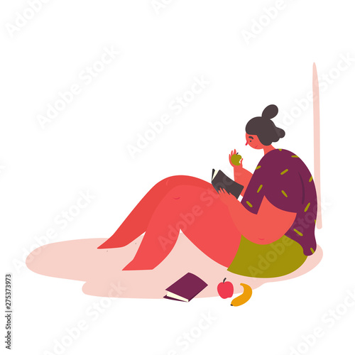 A young woman sits on the floor and reads a book. The girl is resting and eats an apple while reading.