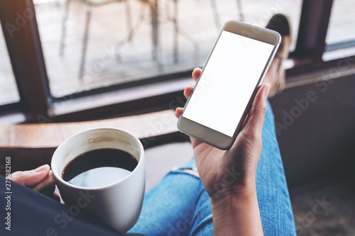 Mockup image of a woman holding white mobile phone with blank desktop screen while sitting and drinking coffee in cafe