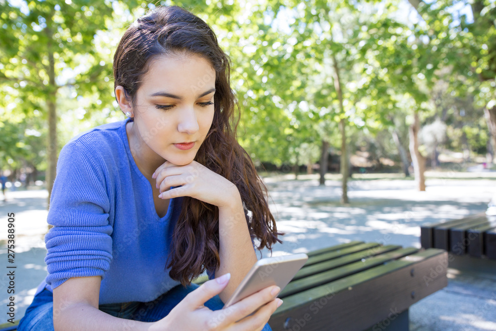 Concentrated woman reading internet blog on smartphone. Serious girl sitting on bench in park and using smartphone. Technology concept