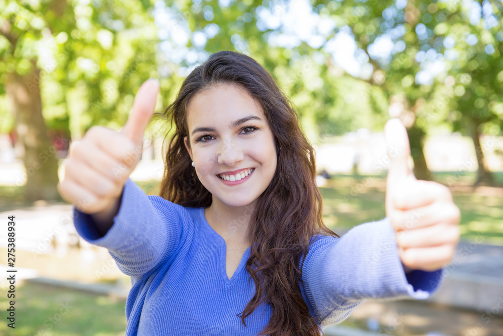 Excited student girl showing thumbs up outdoors. Portrait of successful woman with wavy hair giving estimation using hand sign. Recommending concept Excited student girl showing thumbs up outdoors. Portrait of successful woman with wavy hair giving estimation using hand sign. Recommending concept