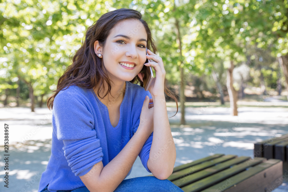 Happy attractive woman talking on cellphone in city park. Portrait of cheerful woman calling friend while sitting on bench in park. Chatterbox concept Happy attractive woman talking on cellphone in city park. Portrait of cheerful woman calling friend while sitting on bench in park. Chatterbox concept