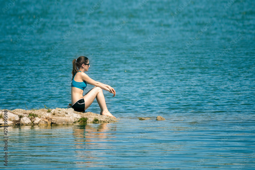 A young girl with sunglasses in a sporty top and black shorts sits thoughtfully on the rocks, looking on a water.