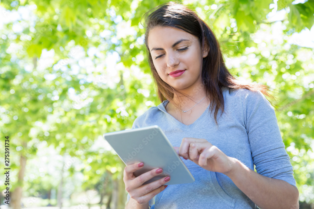Content pretty young lady using tablet computer in park. Beautiful woman wearing hoodie and standing with green trees in background. Technology and nature concept. Front view. Content pretty young lady using tablet computer in park. Beautiful woman wearing hoodie and standing with green trees in background. Technology and nature concept. Front view.