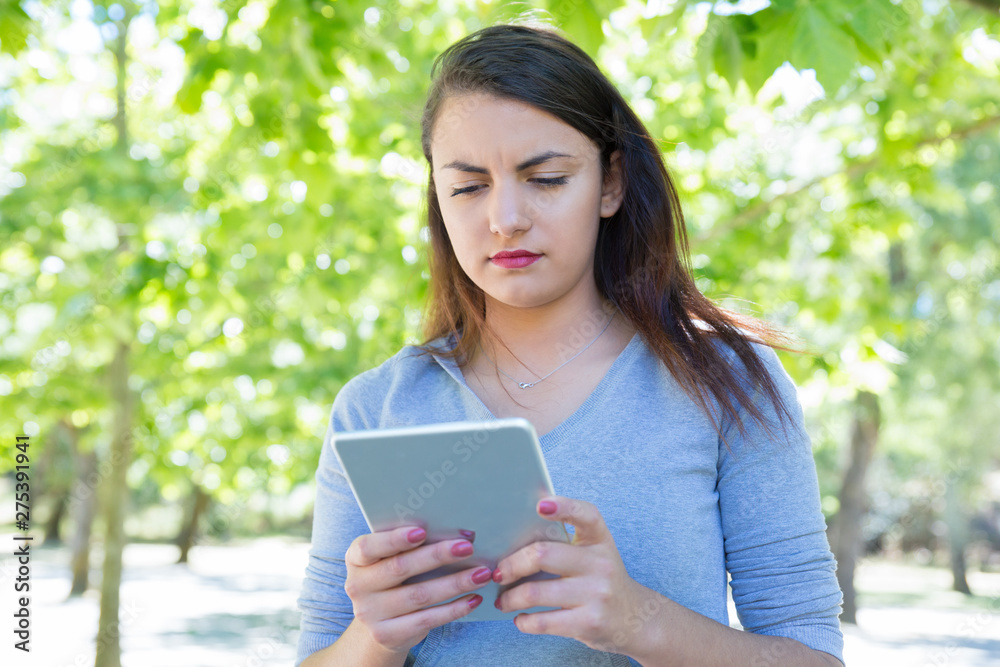 Focused pretty young lady using tablet computer in park. Beautiful woman wearing hoodie and standing with green trees in background. Technology and nature concept. Front view. Focused pretty young lady using tablet computer in park. Beautiful woman wearing hoodie and standing with green trees in background. Technology and nature concept. Front view.