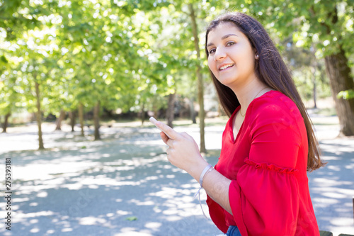 Smiling pretty young lady using smartphone in park. Beautiful woman wearing red blouse and standing with green trees in background. Communication and nature concept.