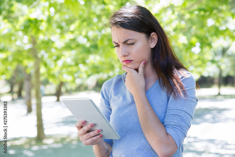 Thoughtful pretty young lady using tablet computer in park. Beautiful woman wearing hoodie, touching chin and standing with green trees in background. Technology and nature concept. Front view.