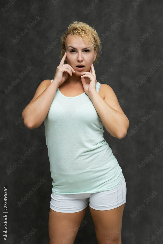 Fototapeta premium Photo portrait above the knee of a beautiful girl blonde woman with short curly hair on a gray background in bright linen is talking and showing a lot of emotions. An experienced model shows hands.
