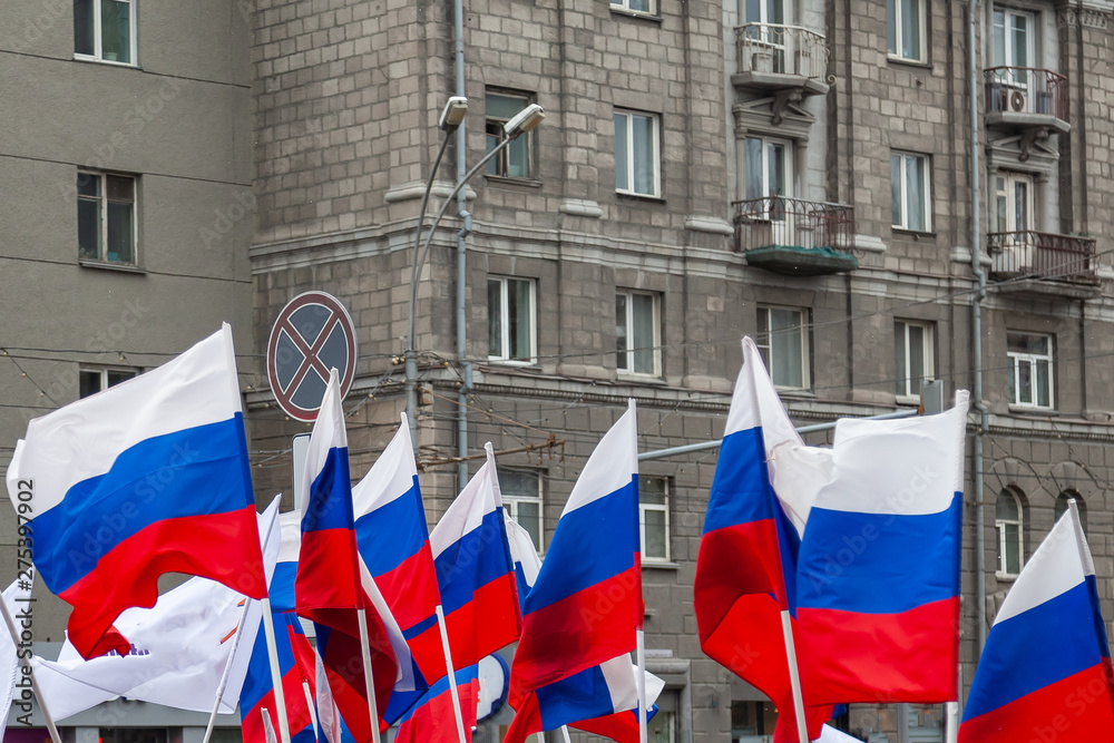 Fototapeta premium A large number of Russian flags in tricolor with stripes of white, blue and red against the background of the walls of buildings with gray windows during a rally or parade