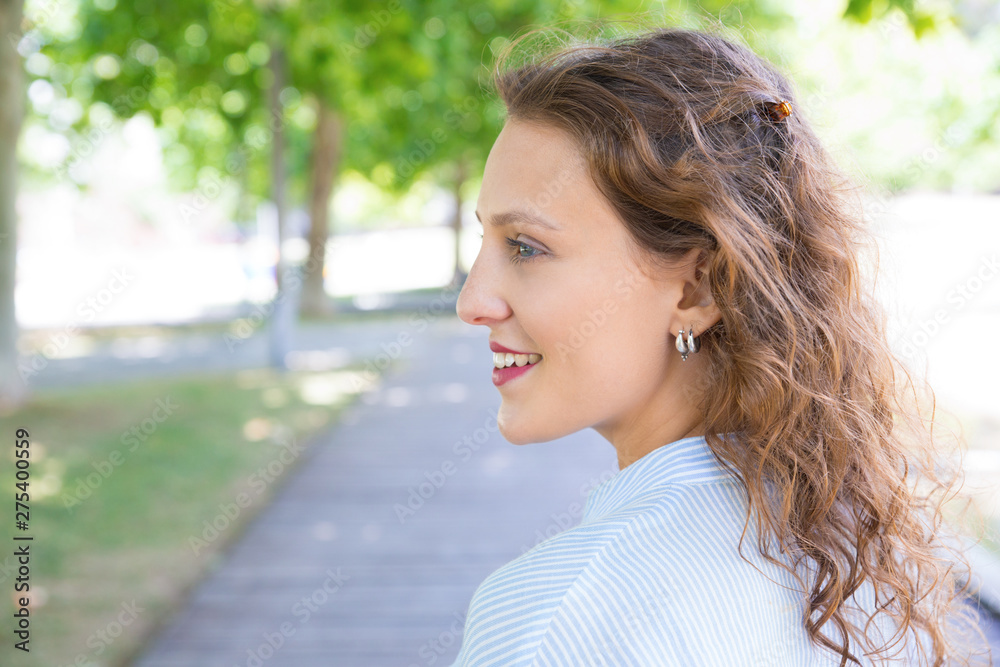 Happy cheerful beautiful girl walking in park. Closeup side view of young woman with wavy hair looking away and smiling. Woman outdoors concept Happy cheerful beautiful girl walking in park. Closeup side view of young woman with wavy hair looking away and smiling. Woman outdoors concept