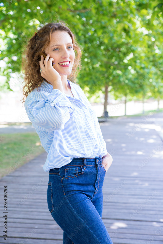 Happy student girl talking on cell outdoors. Beautiful young woman in casual standing on walkway in park, calling on smartphone, listening and smiling. Communication concept
