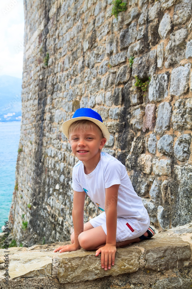 boy in a blue hat sitting on parapet of the ancient fortress of Old town in Budva, Montenegro boy in a blue hat sitting on parapet of the ancient fortress of Old town in Budva, Montenegro