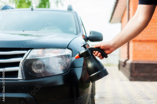 A man sprays car shampoo on a black car from a plastic bottle.