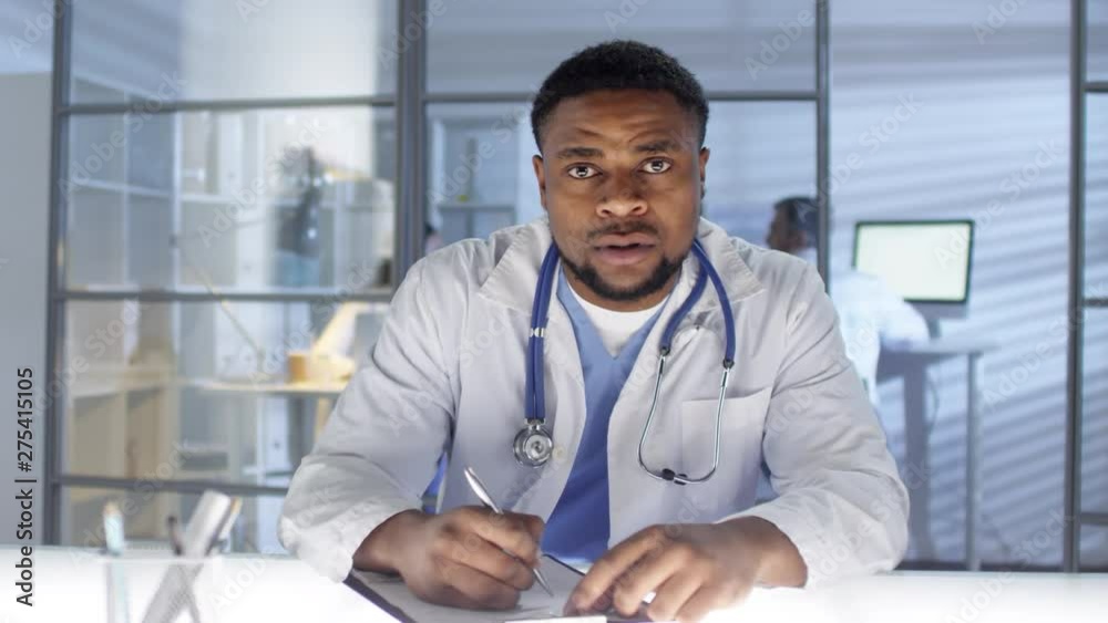 POV shot of attentive and sympathetic middle-aged black GP in white coat at work in his practice, talking to patient during appointment and filling in clinical record with complaints and symptoms POV shot of attentive and sympathetic middle-aged black GP in white coat at work in his practice, talking to patient during appointment and filling in clinical record with complaints and symptoms