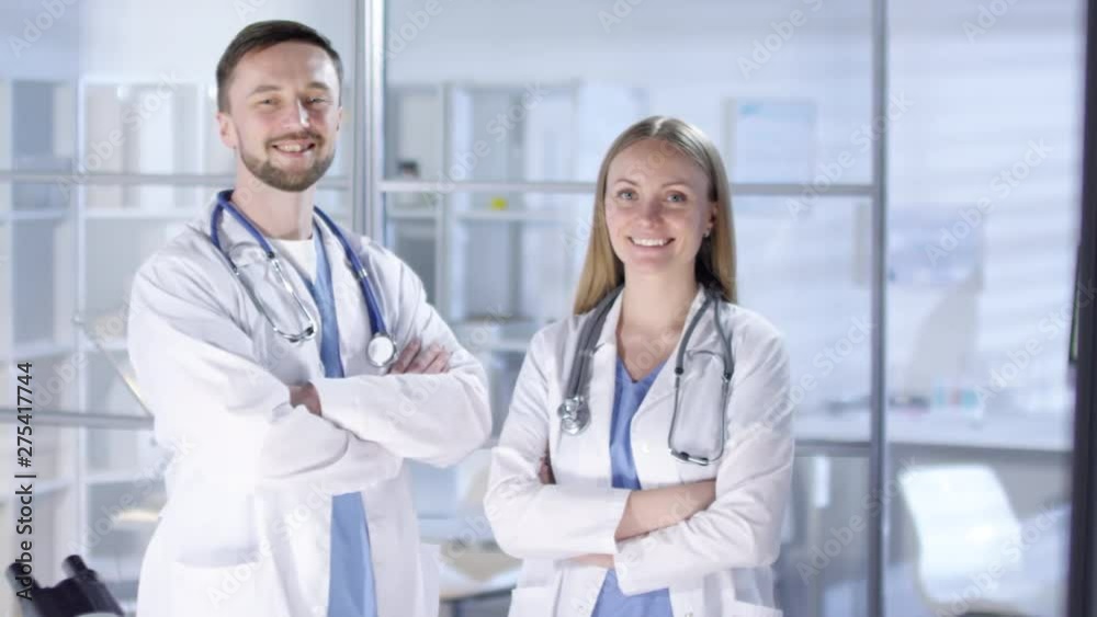 Panning thigh-up shot of Caucasian 30-something male and female physicians at work at hospital, dressed in scrubs and lab coats with stethoscopes, posing together for photos and smiling cheerfully Panning thigh-up shot of Caucasian 30-something male and female physicians at work at hospital, dressed in scrubs and lab coats with stethoscopes, posing together for photos and smiling cheerfully