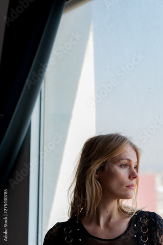 vertical head shot of blond woman looking out of window with sad and melancholic expression below curtain with natural light with clear sky and delicate colors and soft light