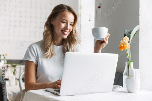Smiling young pretty blonde woman student sitting in cafe using laptop computer drinking coffee.