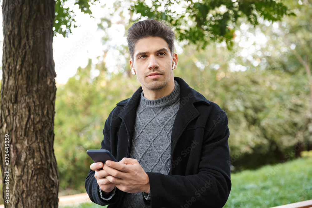 Serious handsome young man in casual clothing walking outdoors in green park using mobile phone listening music with earphones. Serious handsome young man in casual clothing walking outdoors in green park using mobile phone listening music with earphones.