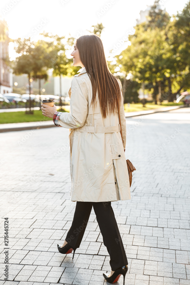 Photo from back of brunette caucasian woman drinking takeaway coffee while walking on city street